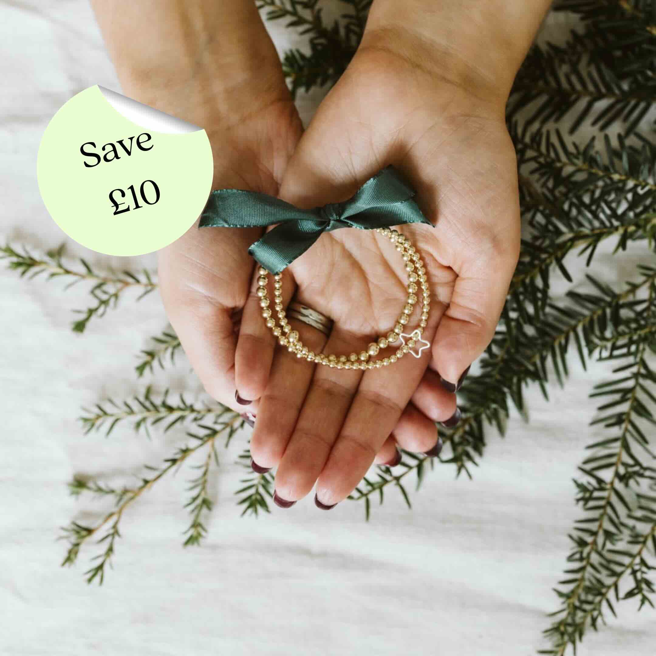 Hands holding gold bracelets with a green bow and 'Save £10' label on a white background with green leaves.