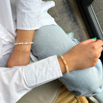 Person wearing a white shirt and light blue jeans with a bracelet on a wooden chair.