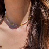 A close-up lifestyle shot of a person wearing a delicate beaded necklace with white, yellow, and pink beads. The necklace is shown worn around the neck, with brown hair and a blue top visible in the background.