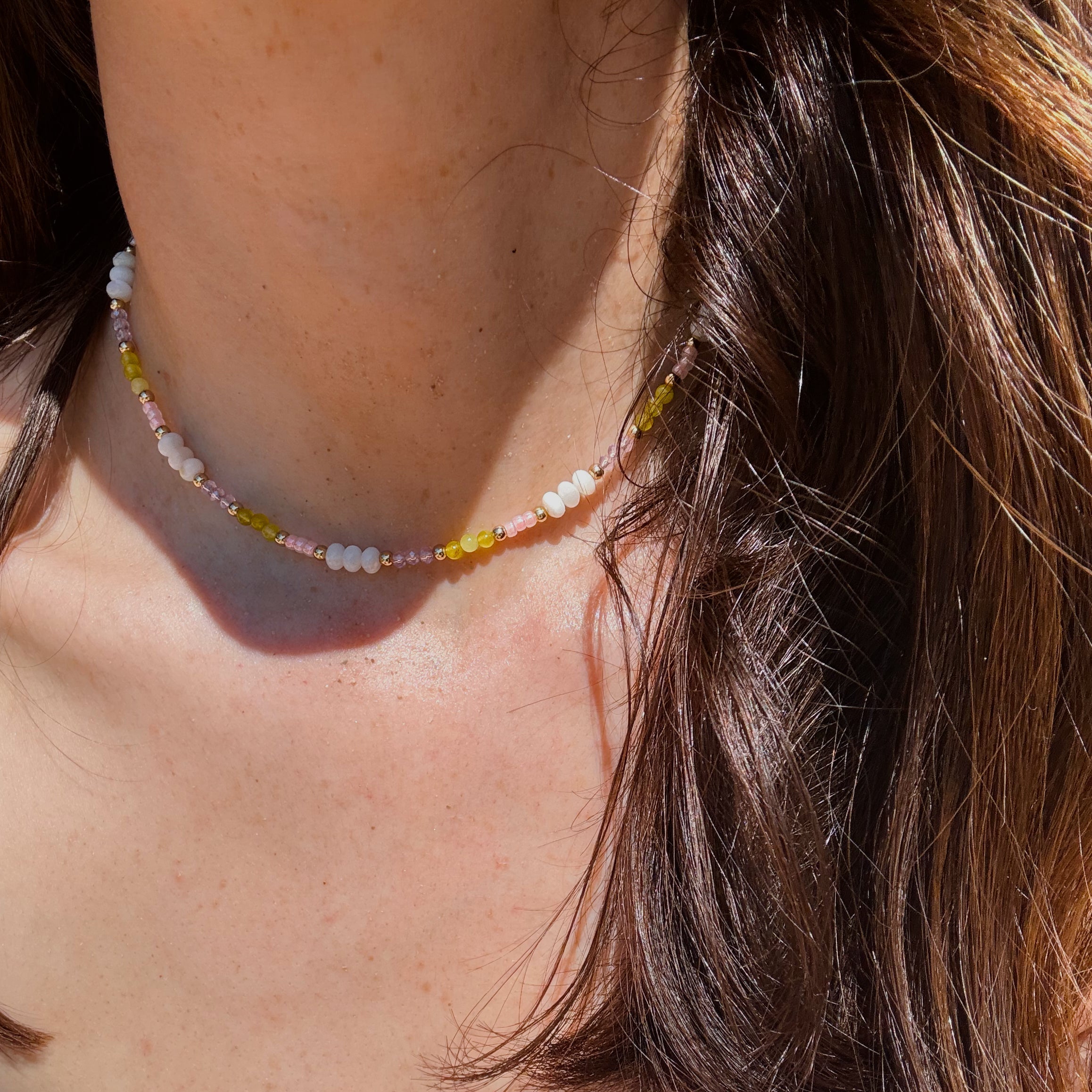 A close-up lifestyle shot of a person wearing a delicate beaded necklace with white, yellow, and pink beads. The necklace is shown worn around the neck, with brown hair and a blue top visible in the background.