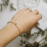 A close-up of a person's hand wearing a delicate gold beaded bracelet with a tiny white pearl accent, styled with gold rings and a lace trinket dish on a white surface with pine branches.