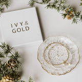 A styled flat-lay featuring a white jewelry box with the brand name 'IVY & GOLD' in gold lettering, alongside a scalloped white dish with gold trim holding delicate gold bracelets, surrounded by pine branches and pinecones on a white background.