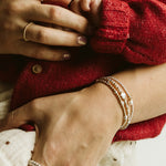 Close-up lifestyle shot of a baby in a red knit sweater being held by an adult, showcasing delicate gold jewelry including rings and bracelets on the adult's fingers and wrist.