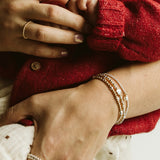 Close-up lifestyle shot of a baby in a red knit sweater being held by an adult, showcasing delicate gold jewelry including rings and bracelets on the adult's fingers and wrist.