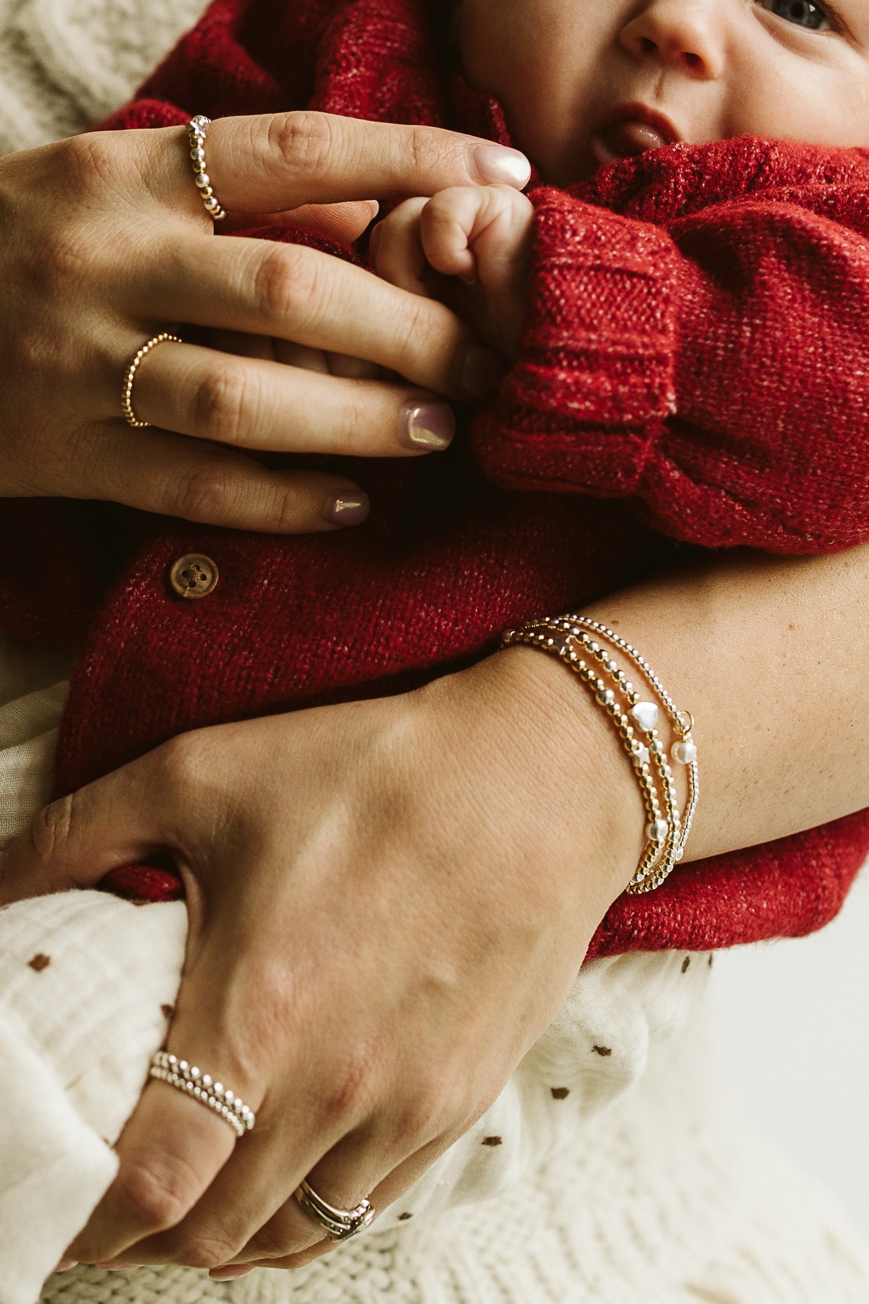 Close-up lifestyle shot of a baby in a red knit sweater being held by an adult, showcasing delicate gold jewelry including rings and bracelets on the adult's fingers and wrist.