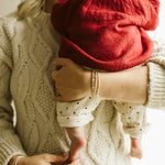 A close-up lifestyle shot of a woman in a cream cable-knit sweater holding a baby dressed in a red knit bonnet and cream polka-dot pants.