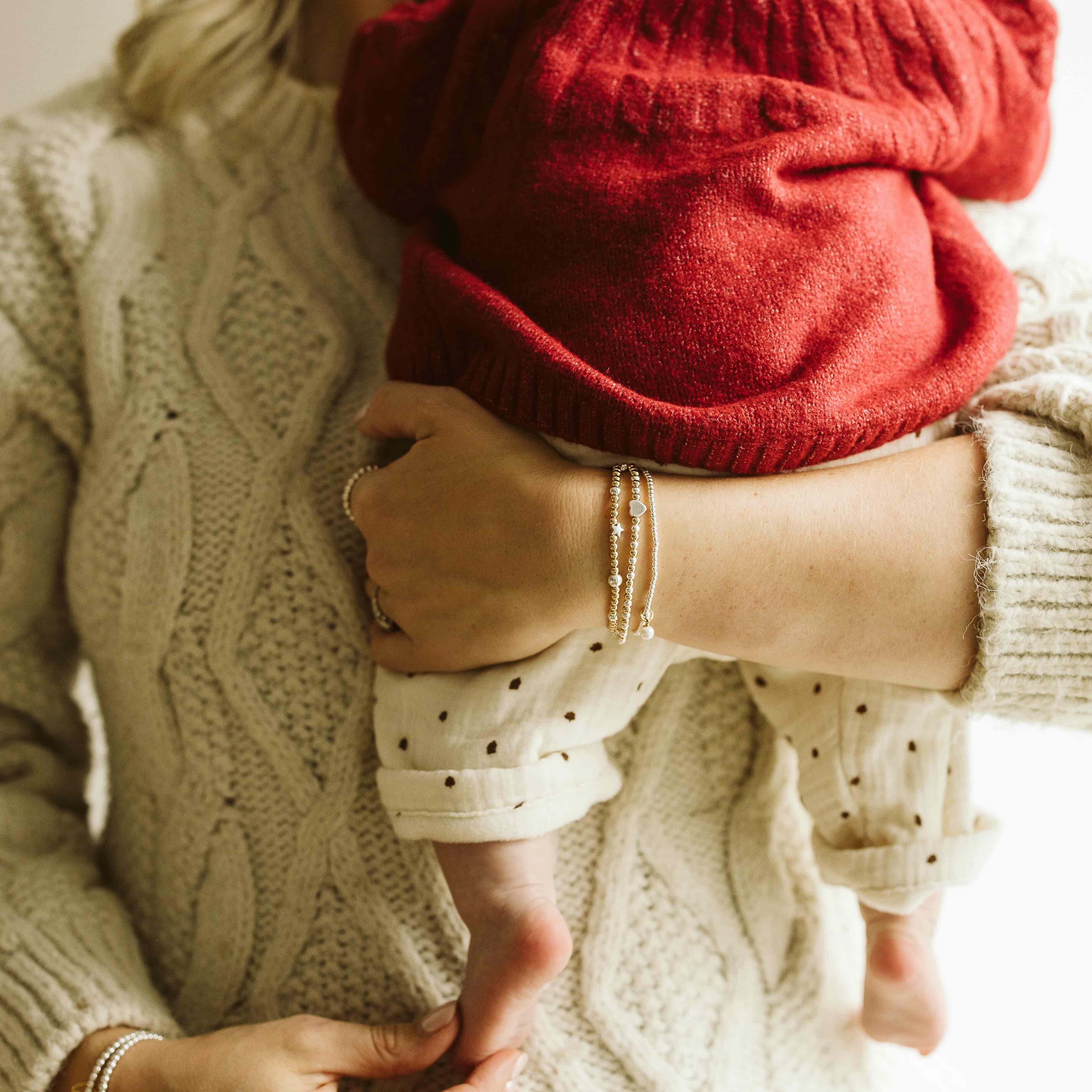A close-up lifestyle shot of a woman in a cream cable-knit sweater holding a baby dressed in a red knit bonnet and cream polka-dot pants.